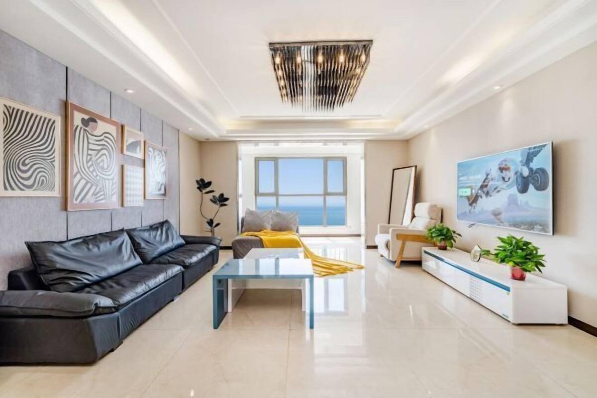 A modern living room with a black leather sofa, a glass coffee table, and a white console table with plants. There are abstract wall art pieces and a large framed picture on the wall.