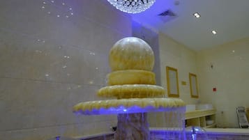 A marble fountain with a spherical top and a base, located in a room with a tiled wall and a reception area in the background.