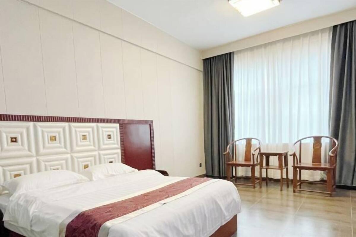 A hotel room with a large bed, a wooden headboard, a small table with chairs, and a window with curtains.