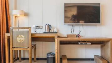 A compact hotel room with a wooden desk, a television mounted on the wall, a lamp, and a trash bin.