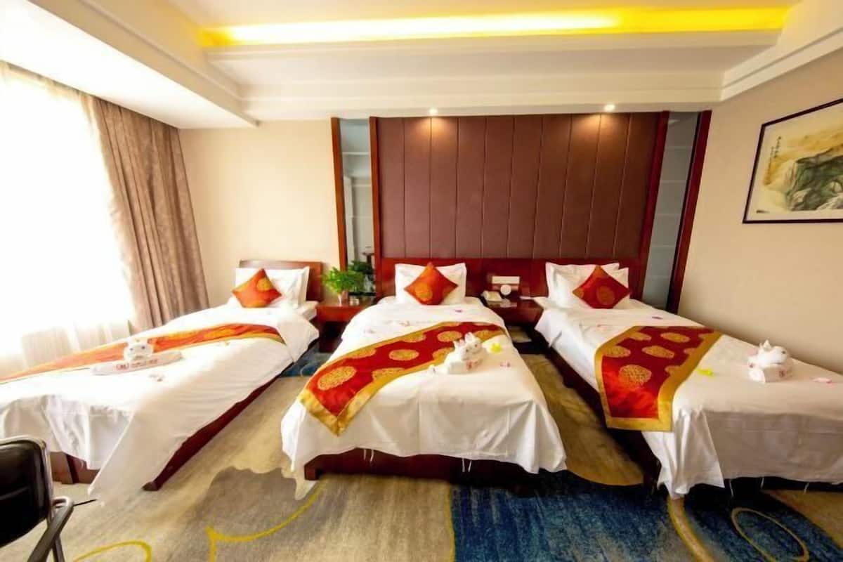 Habitación de hotel con tres camas, cada una con ropa de cama blanca y detalles rojos, cabeceros de madera y un cuadro en la pared.