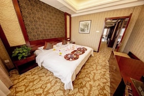 A hotel room with a large bed, a nightstand, a dresser, and a plant. - Han Tang Grand Hotel (Jiuquan)