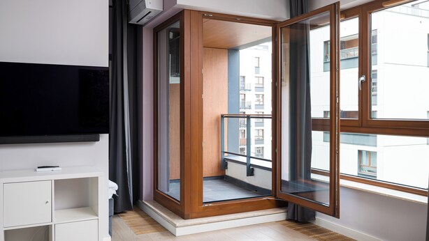 Un apartamento moderno con un amplio ventanal corredera que da a un balcón, un televisor de pantalla plana montado en la pared y un mueble blanco de almacenamiento.