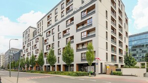 Un moderno edificio residencial de varios pisos con balcones, entrada vigilada y un área ajardinada con árboles.