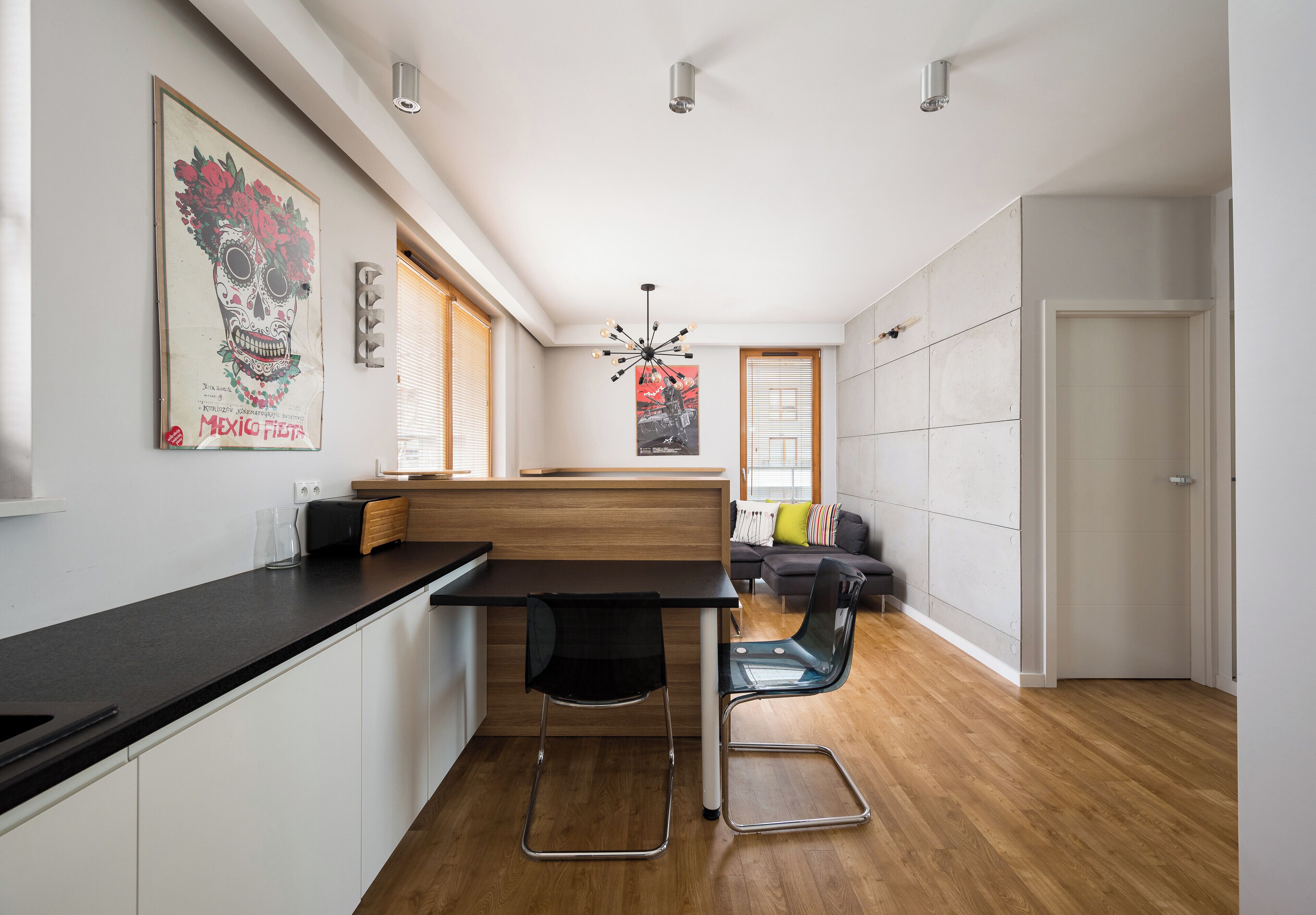 Een moderne keuken met een zwart aanrecht, houten kastjes en een zwarte stoel. Er staat een houten eettafel met een zwarte stoel. De kamer heeft een houten vloer, witte muren en een plafondlamp.