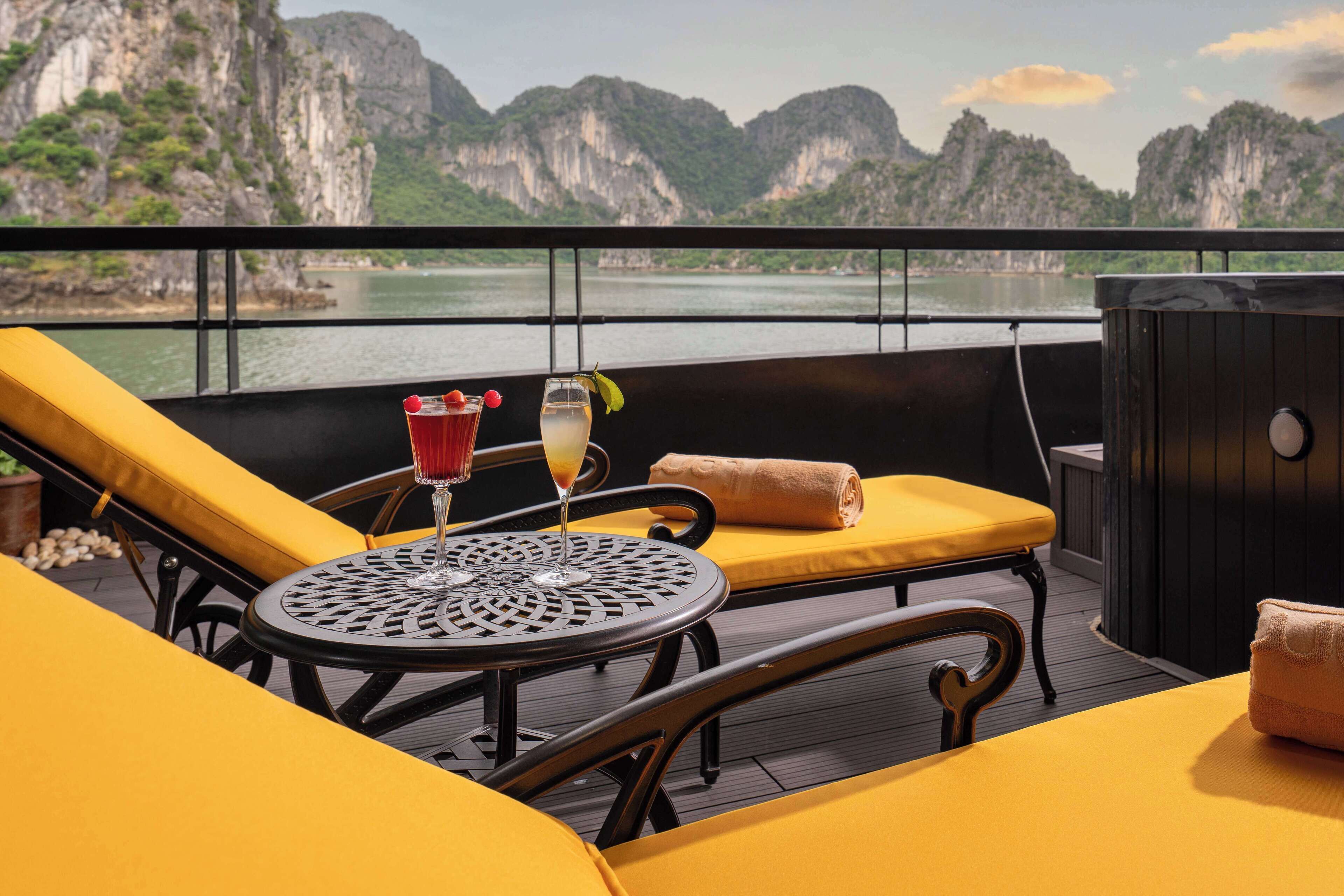 Un balcon avec des sièges rembourrés de couleur jaune, une table ronde avec une boisson, et une vue sur une rivière et des montagnes.