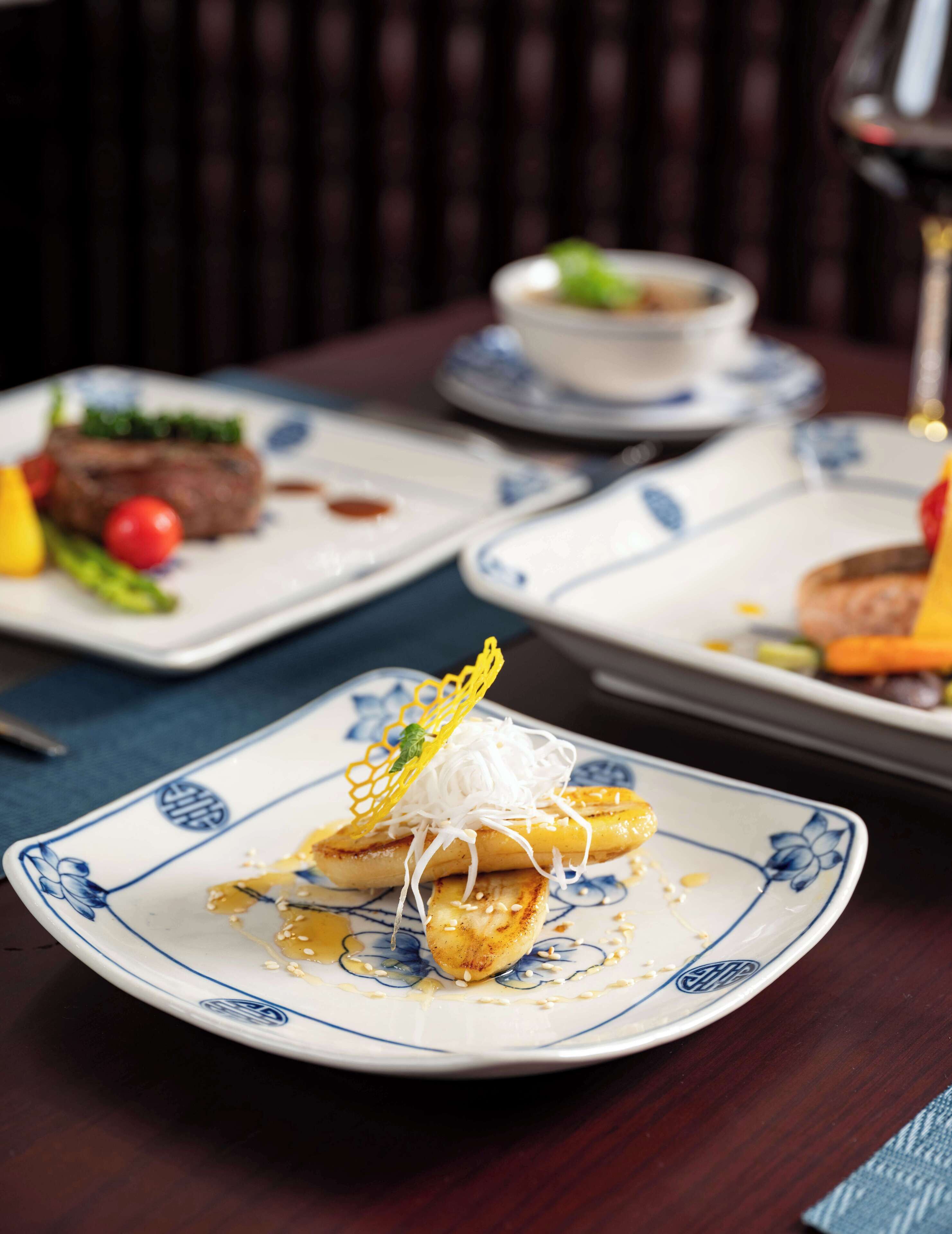 A dining table with plates of food, including a dish with banana slices and white noodles, and another with a steak and vegetables.