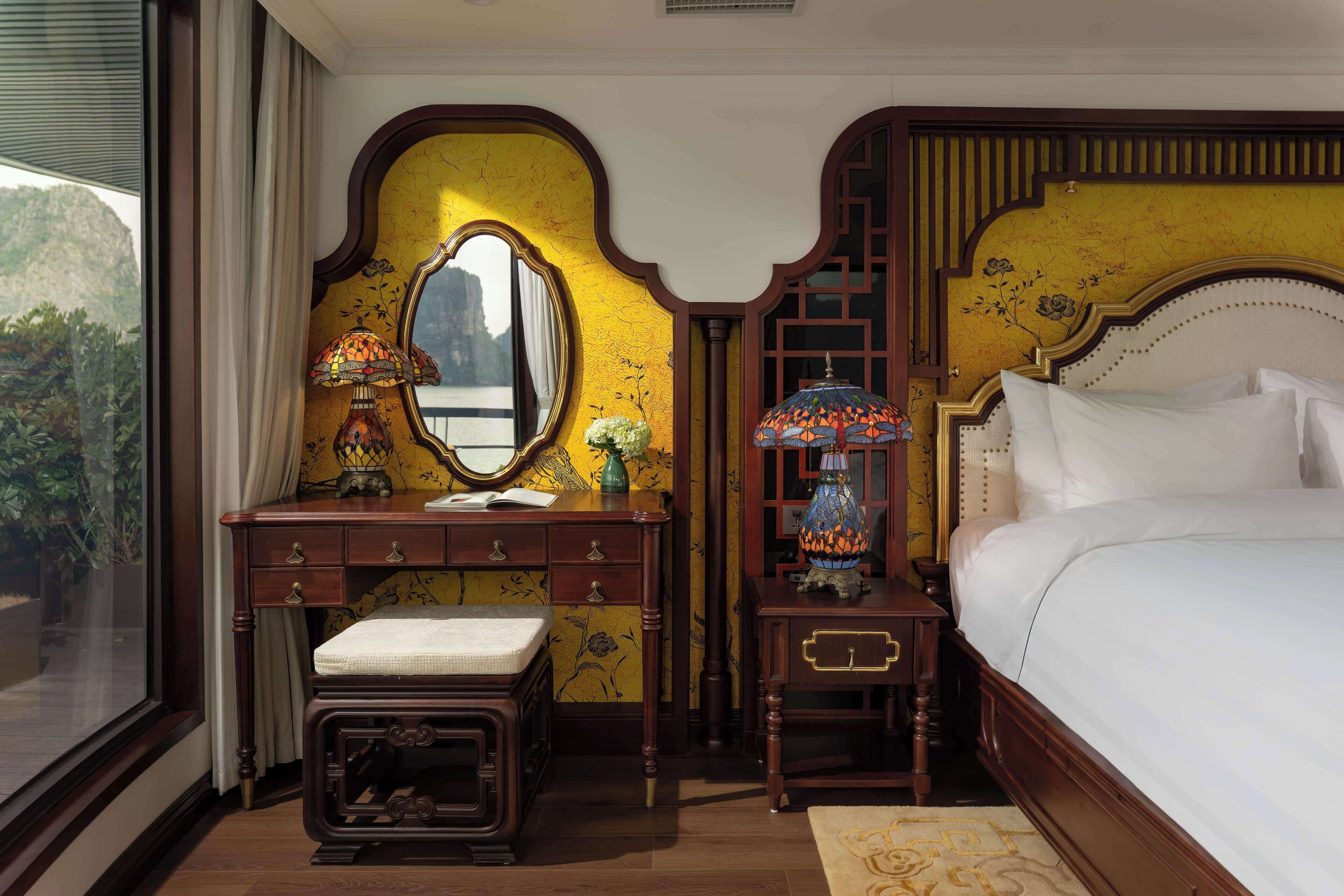Habitación de hotel con una cama grande, un aparador de madera con cajones, un espejo y una mesita de noche con lámpara.