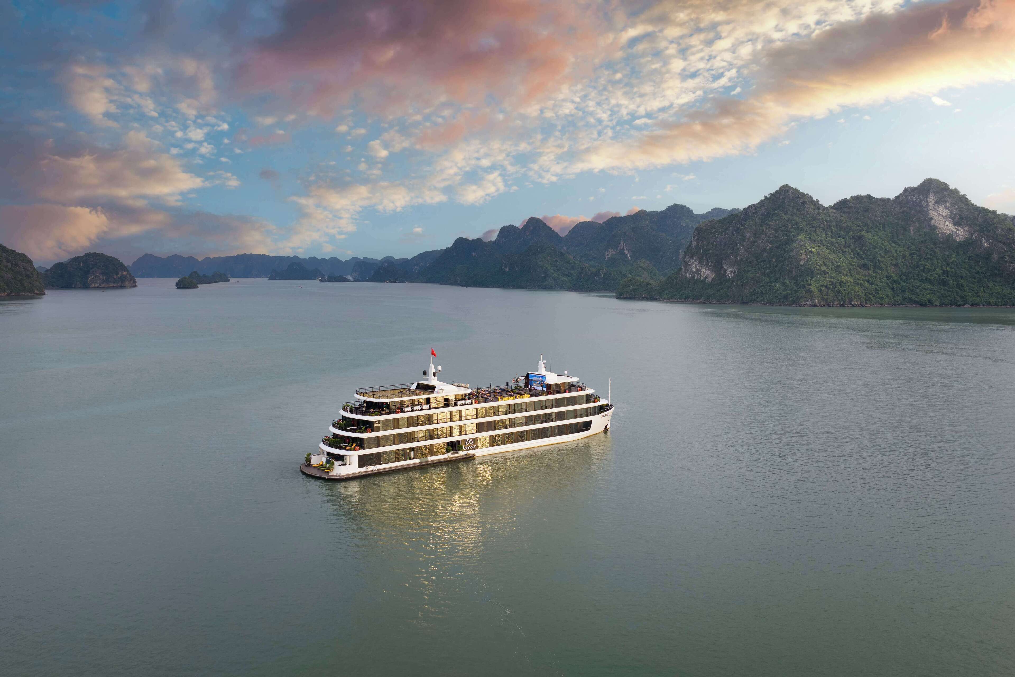 Een groot, meerdekker cruiseschip met meerdere balkons, vaart over rustig water met een achtergrond van bergachtige eilanden en een gedeeltelijk bewolkte lucht.