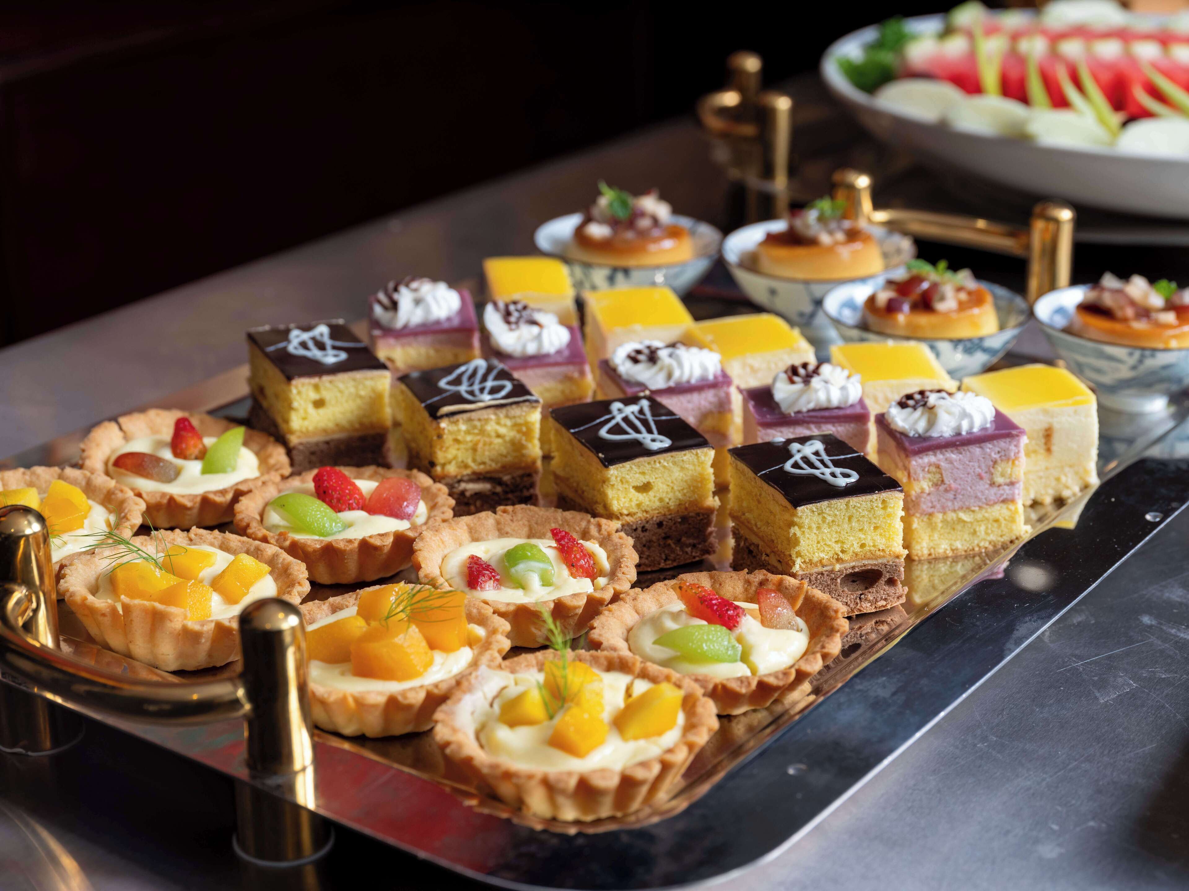 A variety of desserts on a tray, including tarts and layered cakes, with some garnished with fruits and cream.