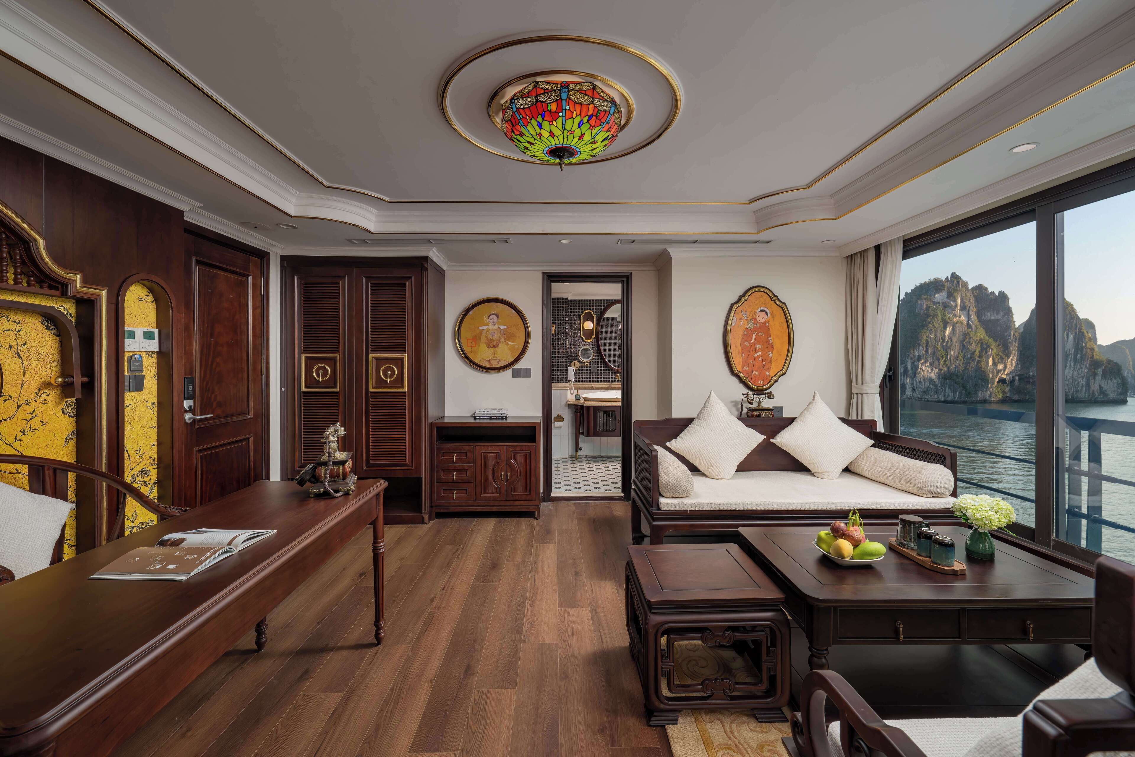 A spacious room with a wooden floor, a white sofa with cushions, a coffee table with fruit, and a large window offering a view of a body of water and cliffs.
