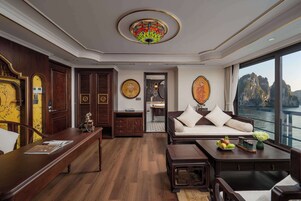 Amplia habitación con piso de madera, un sofá blanco con cojines, una mesa de centro con fruta y un gran ventanal con vistas al agua y acantilados.
