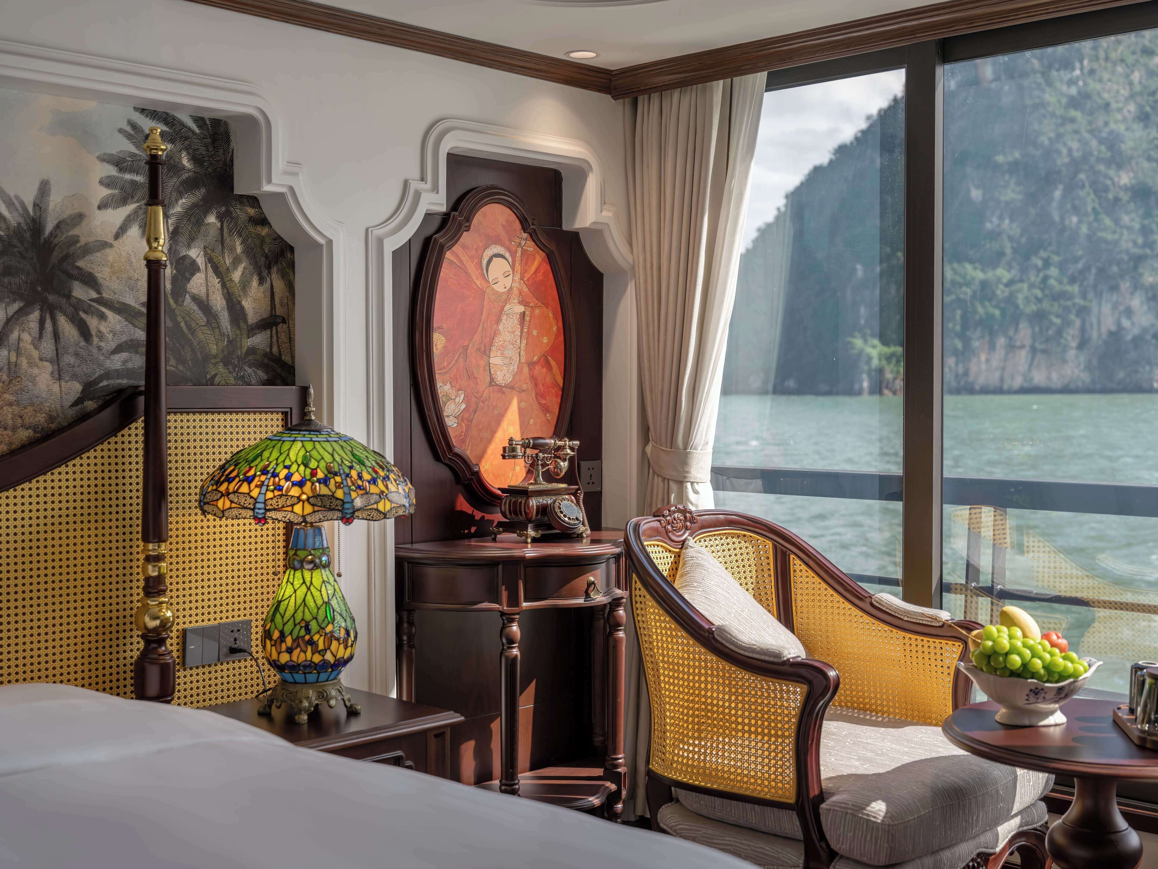 A hotel room with a bed, a chair, a table with fruit, a decorative lamp, and a painting.