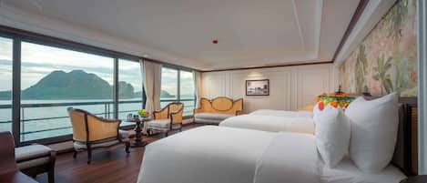 Habitación de hotel con dos camas, zona de estar con sillas, una mesa con frutero y un ventanal con vistas al mar y las montañas.
