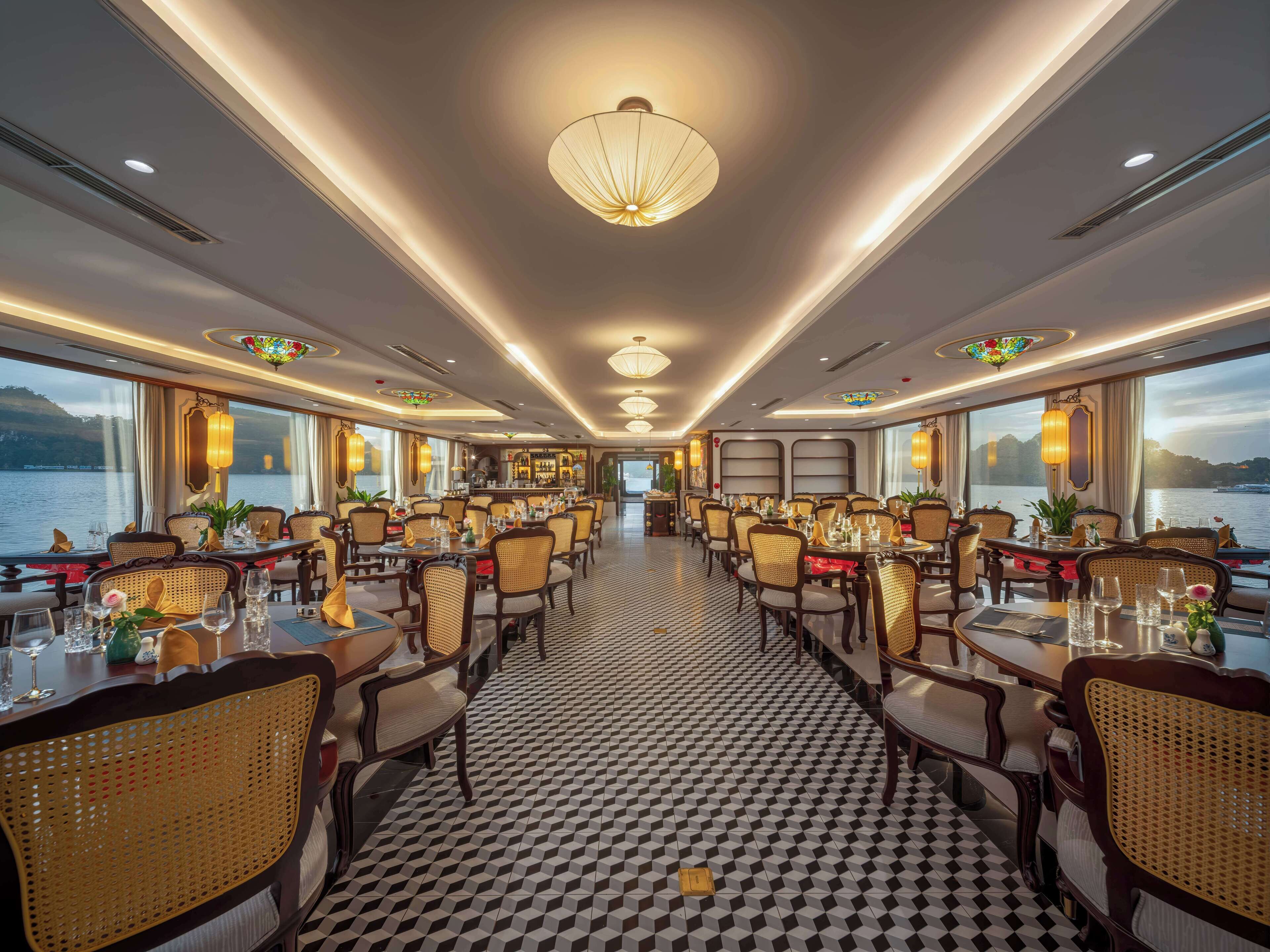 A spacious dining area with multiple tables and chairs, large windows offering views of the water, and a patterned floor.