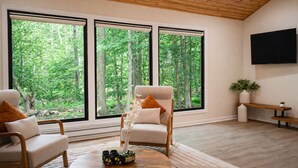 Un salon doté d’une grande fenêtre donnant sur une forêt, d’un plafond en bois et d’un téléviseur à écran plat fixé au mur.