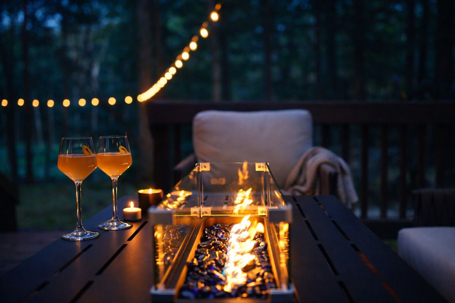 A wooden deck with a fire pit, two glasses of a beverage, and string lights in a forested area.