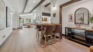 A spacious dining area with a long wooden table, several chairs, and a modern kitchen in the background. - Spacious 7-Bed Home w/ Hot Tub, Fireplace, Gym, Wine Bar, and 2 Living Spaces! (Lakeside)