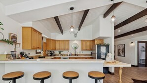 A modern kitchen with wooden cabinets, a central island with stools, pendant lighting, and a rustic ceiling. - Spacious 7-Bed Home w/ Hot Tub, Fireplace, Gym, Wine Bar, and 2 Living Spaces! (Lakeside)