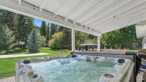 A hot tub under a covered patio with a view of a lawn and trees. - Spacious 7-Bed Home w/ Hot Tub, Fireplace, Gym, Wine Bar, and 2 Living Spaces! (Lakeside)