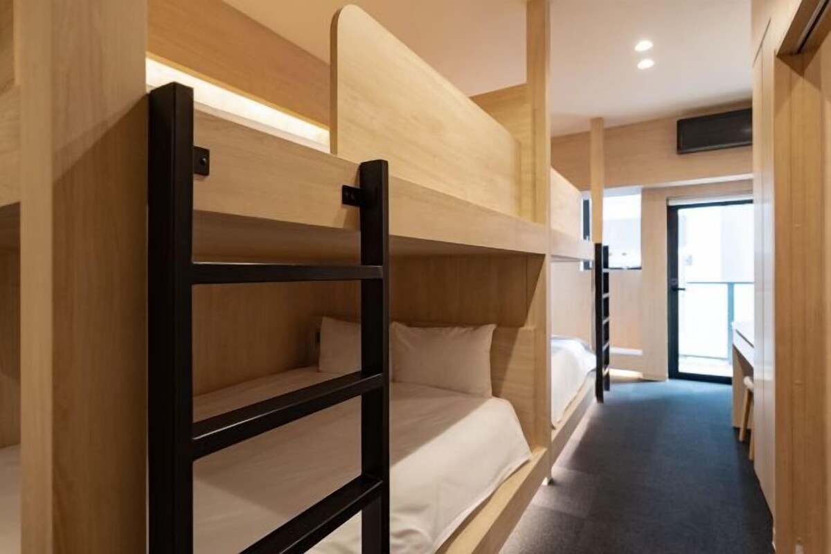 Une chambre avec un lit superposé, une tête de lit en bois, un petit bureau et une fenêtre laissant entrer la lumière naturelle.