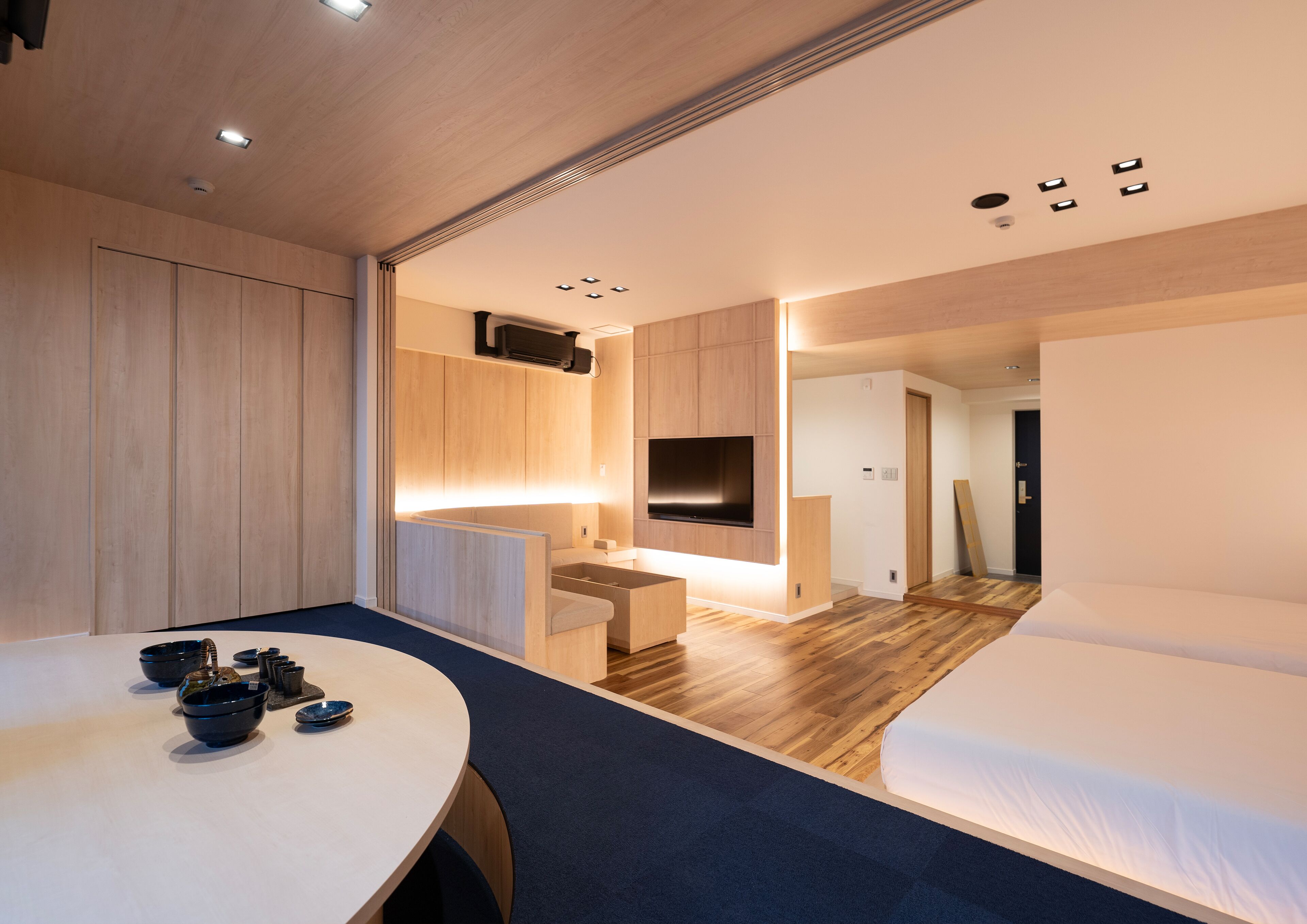 A modern hotel room with a wooden floor, a bed, a desk with a flat-screen TV, and a seating area with a table and chairs.