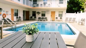 Un espace piscine agrémenté d’une table en bois et d’une plante en pot, entouré de chaises longues et d’un bâtiment doté de balcons.