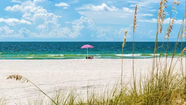 Une plage où une personne se détend sous un parasol rose, avec du sable blanc et de l’herbe haute au premier plan.