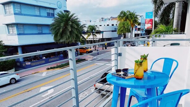 A balcony with blue chairs and a table, overlooking a street with cars and buildings. - Comfortable SoBe Studio | Balcony | Free Beach Chairs & More (Miami Beach)