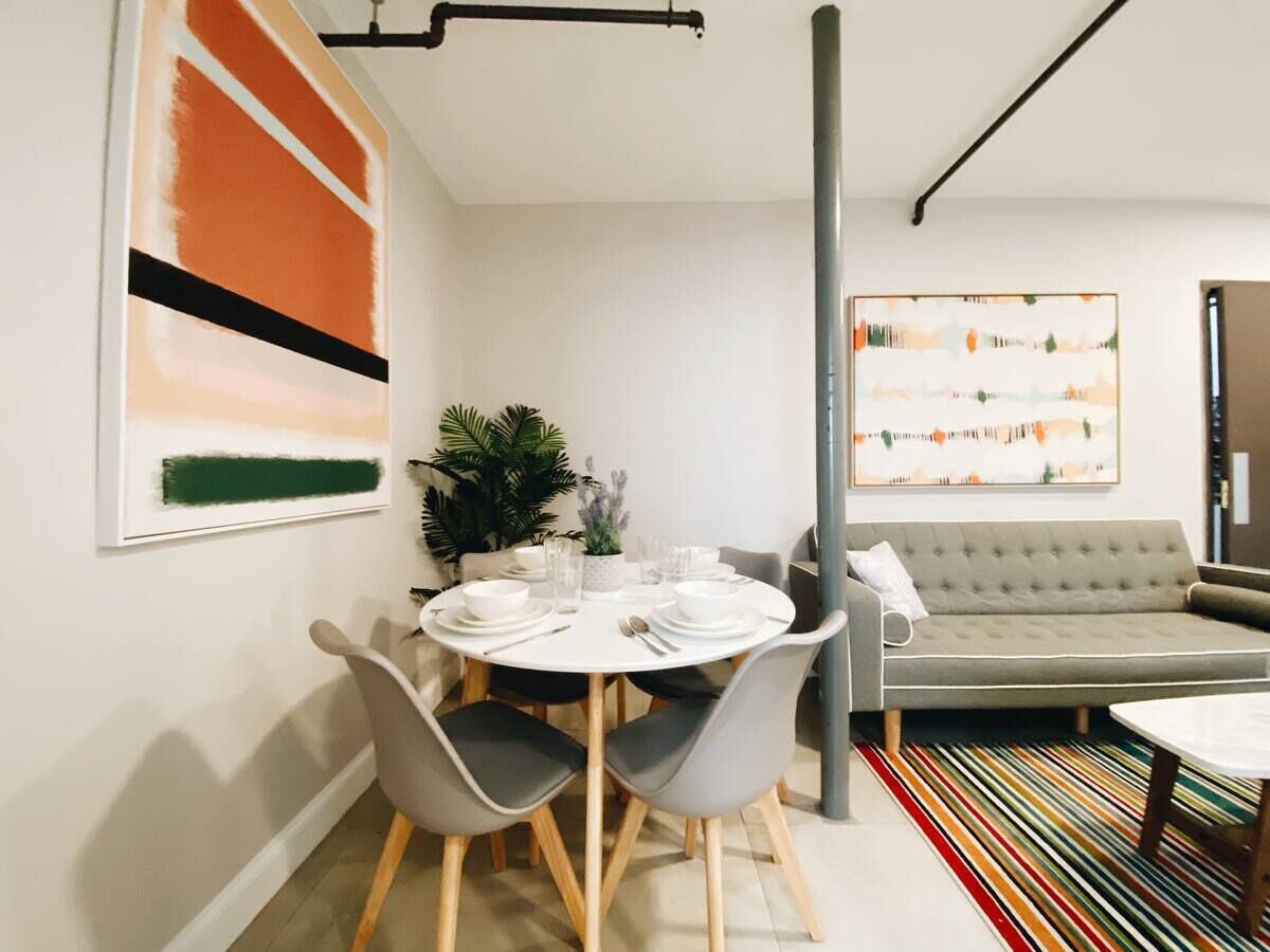 Ein moderner Essbereich mit einem runden Tisch, gedeckt für vier Personen, umgeben von grauen Stühlen. Im Hintergrund sind eine graue Couch und ein farbenfroher Teppich mit Streifen zu sehen.