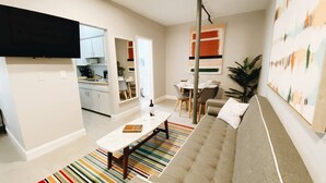 A modern living room with a sofa, a coffee table, and a striped rug. There is a kitchen area in the background with cabinets and a dining table.