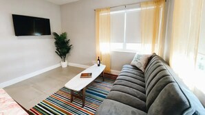A living room with a sofa, a coffee table, a TV mounted on the wall, and a window with curtains. - Spacious Unit | Free Beach Chairs | Near Ocean (Miami Beach)