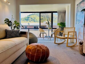 Ein modernes Wohnzimmer mit einer Couch, einem Hocker und Korbstühlen. Ein großes Fenster bietet Blick auf eine gebirgige Landschaft.