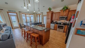 A spacious kitchen with wooden cabinets, a central island with stools, a microwave, oven, and a refrigerator. - Accessible Semi-Soundfront: Pool, Hot Tub, Game Rm, Elevator, Boat Ramp, Linens (Rodanthe)