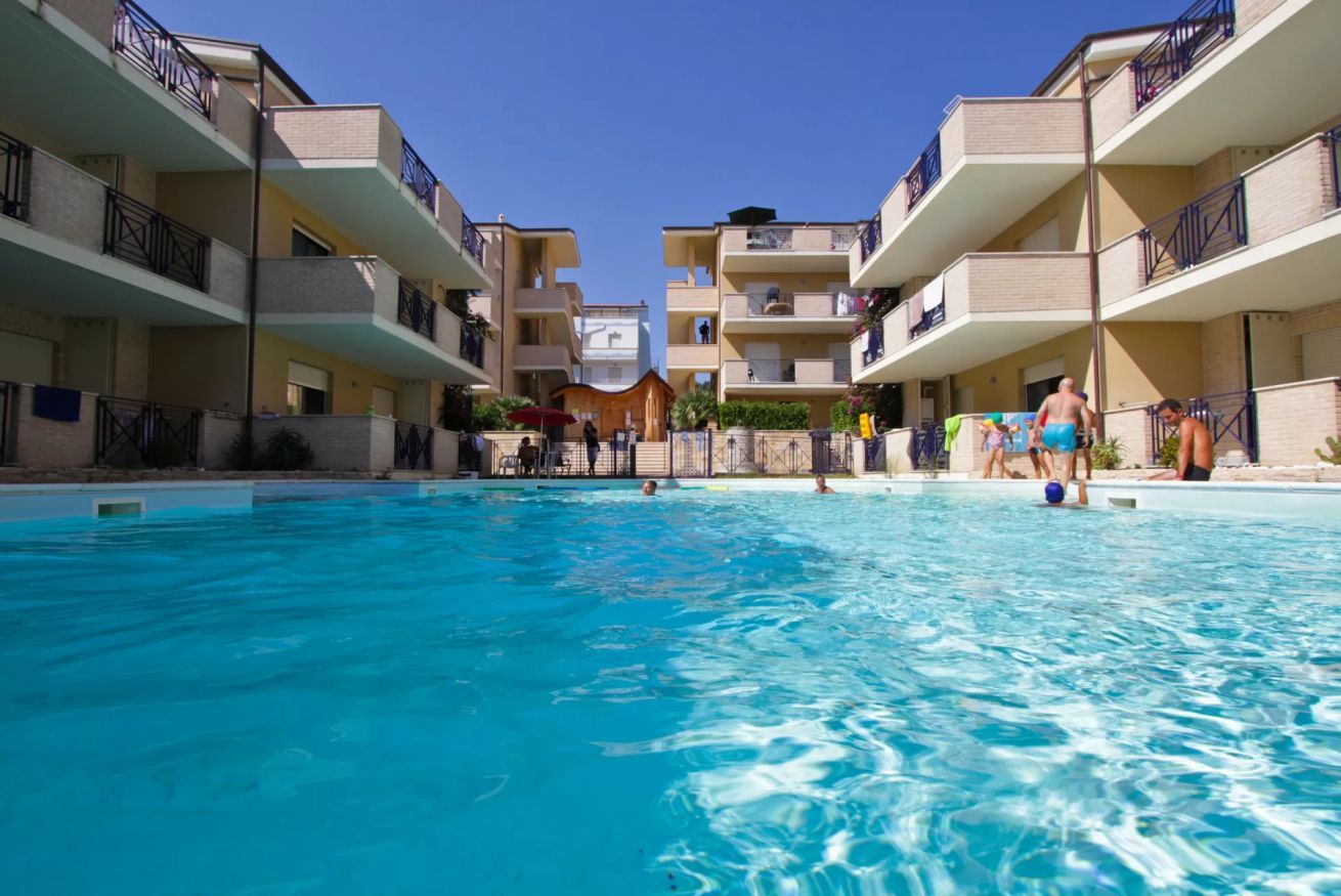 Une piscine entourée d’immeubles à plusieurs étages dotés de balcons, avec une eau d’un bleu limpide et quelques personnes qui nagent.