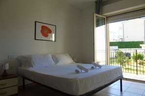 Habitación de hotel con cama, mesita de noche, un cuadro en la pared y balcón con vistas a zonas verdes y edificios.