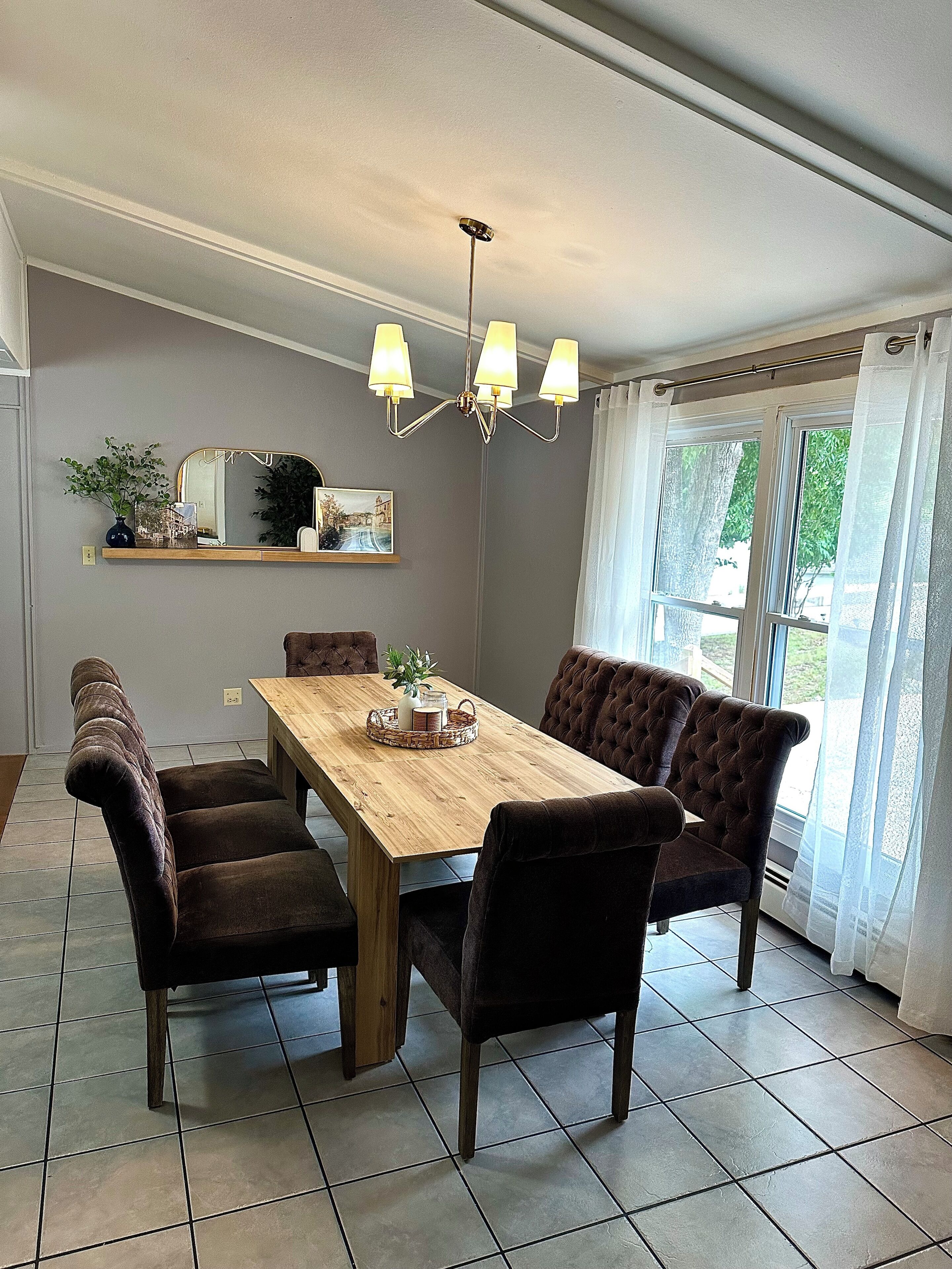 A dining room with a wooden table, six upholstered chairs, a chandelier, a mirror, and a window with curtains.