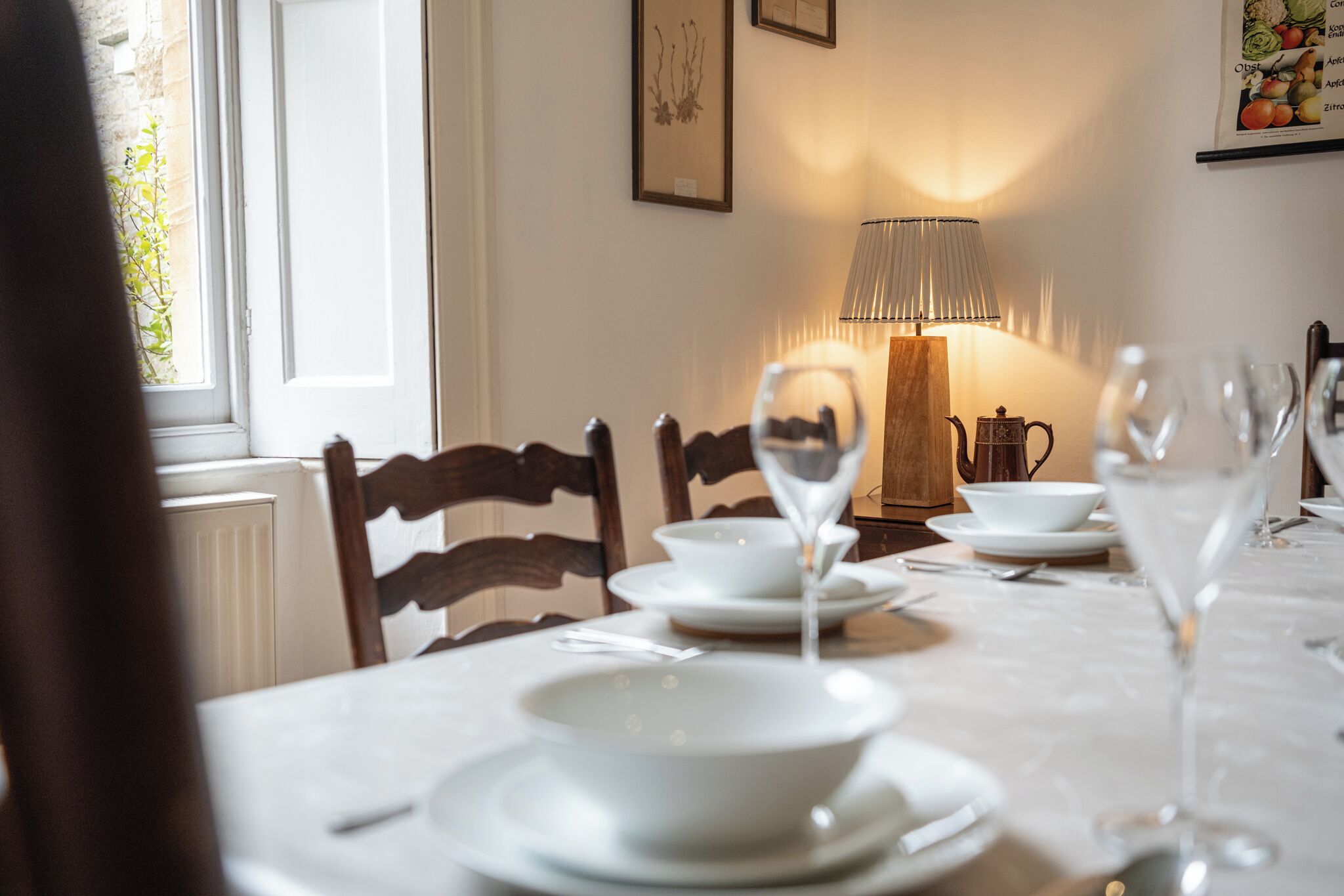 Et spisebord dækket med hvide tallerkner, skåle og vinglas, en lampe samt en tekedel.