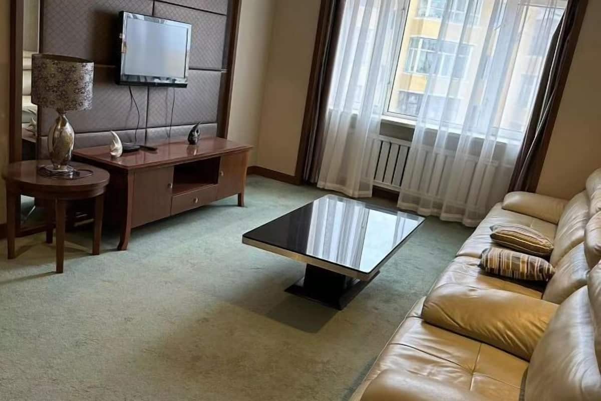 A living room with a sofa, a coffee table, a TV mounted on the wall, and a window with curtains.