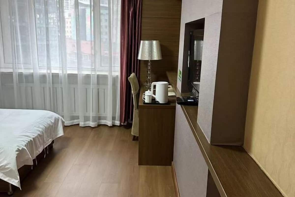 A hotel room with a bed, a desk with a lamp, a coffee maker, and a view of buildings through sheer curtains.