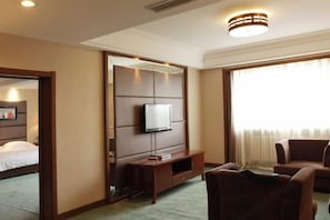 Une chambre d’hôtel comprenant un lit, un téléviseur fixé au mur, un coin salon avec un canapé et des fauteuils, et une grande fenêtre avec des rideaux.