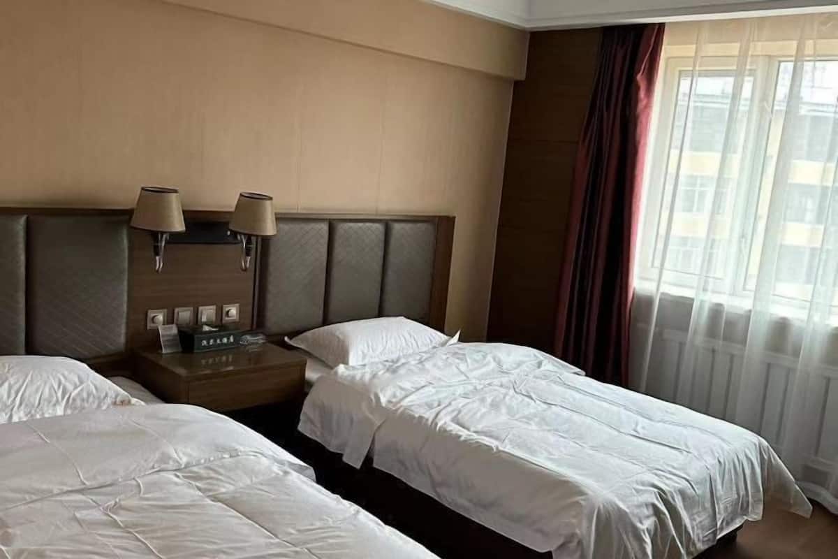 A hotel room with two beds, a wooden headboard, a nightstand with lamps, and a window with curtains.