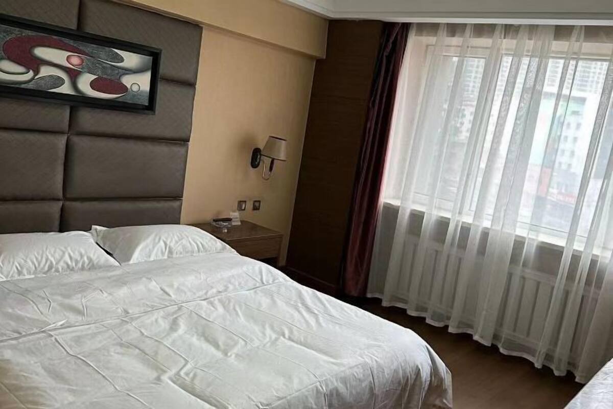 A hotel room with a large bed, a bedside table with a lamp, and a window with sheer curtains.