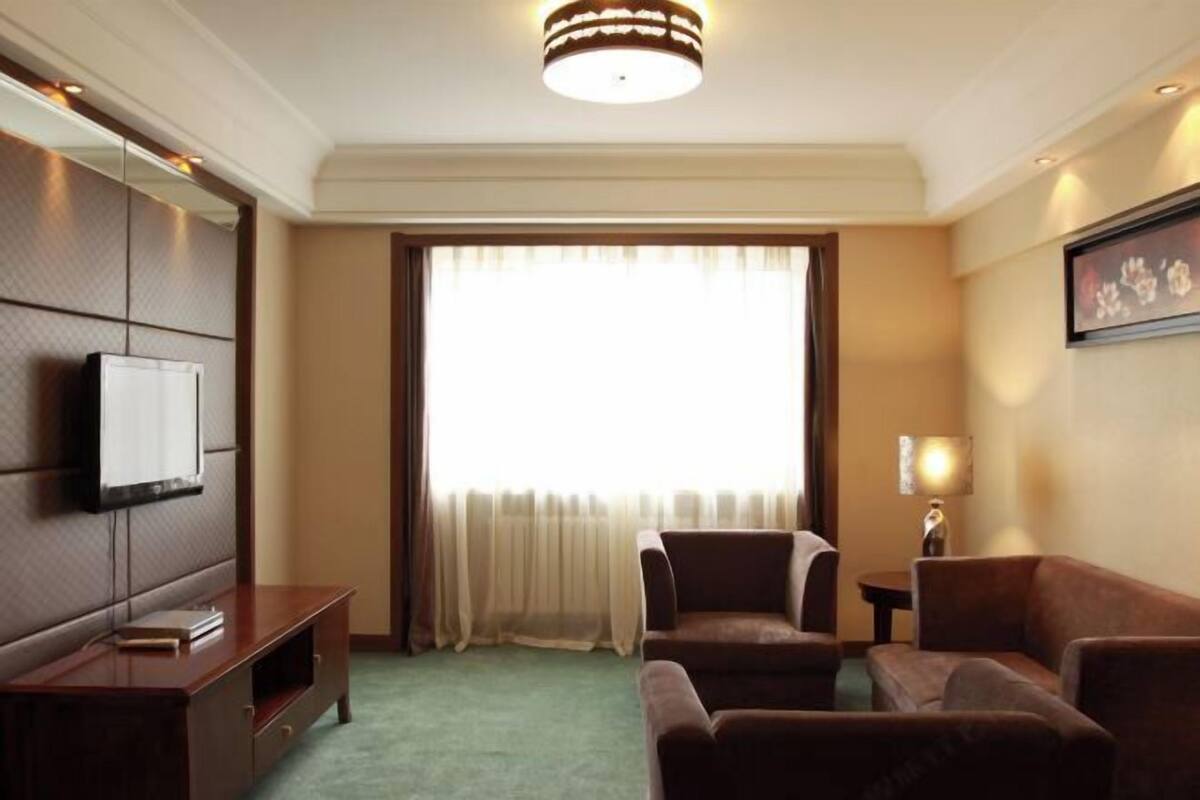 A hotel room with a large window, a flat-screen TV mounted on the wall, a sofa, a small table, and a lamp.