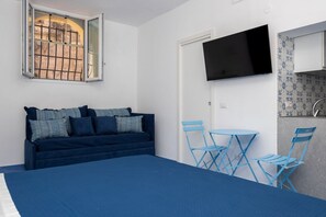 Ein Schlafzimmer mit Bett, einem Fenster mit Metallgitter, einem an der Wand montierten Fernseher und einer kleinen Essecke mit blauen Stühlen und einem Tisch.
