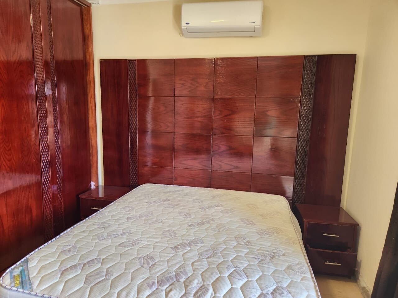 Un dormitorio con cabecera de madera, colchón, mesita de noche y aire acondicionado instalado en la pared.