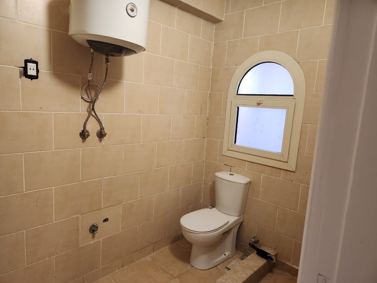 A small, tiled bathroom with a toilet, a window, and a water heater.