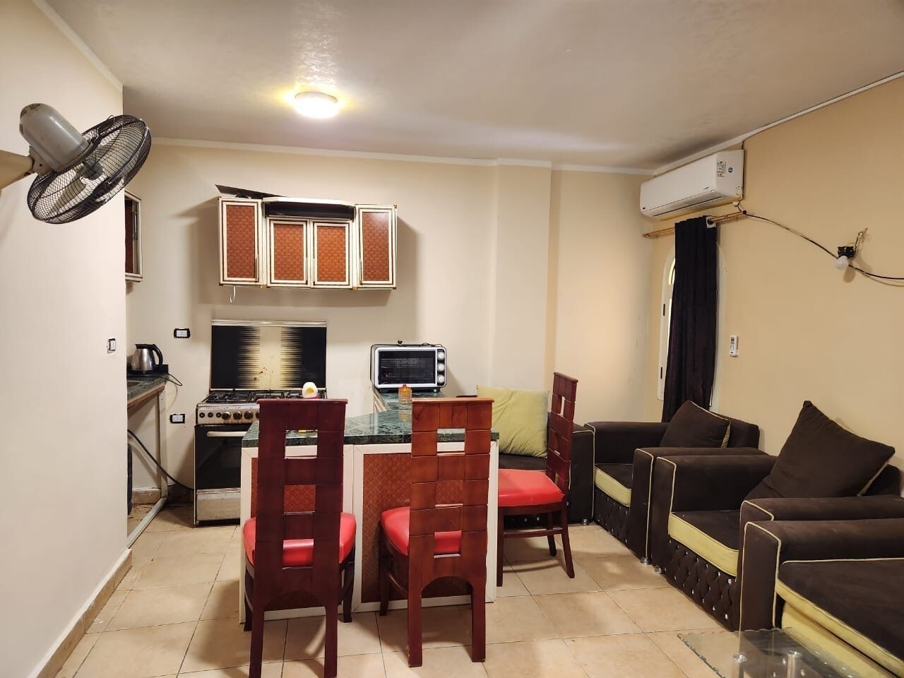 Un espace de vie compact comprenant un coin cuisine, un coin repas avec des chaises en bois, un canapé et un ventilateur de plafond.