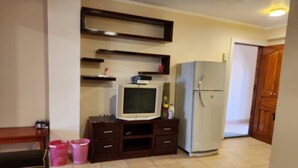 Una cocina compacta con refrigerador, microondas y un televisor pequeño sobre un mueble de madera. Hay dos cestas rosas en el suelo.