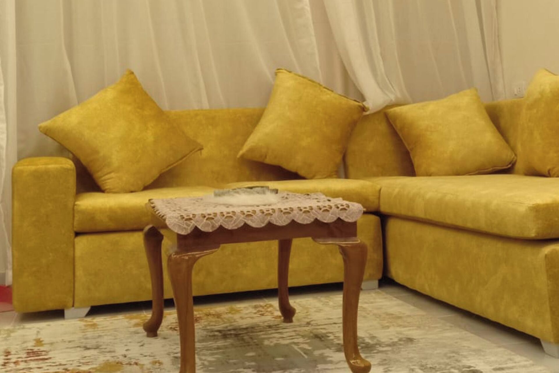 Ein gelbes Sofa mit passenden Kissen, ein kleiner Holztisch mit Spitzentuch und ein weißer Vorhang im Hintergrund.